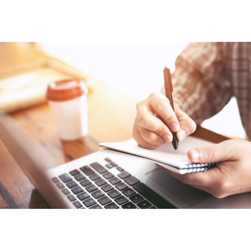 Person writing in a notebook in front of a laptop with a coffee cup