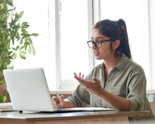 Person with ponytail and glasses speaking to laptop screen and taking notes