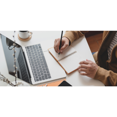 Person Writing on Notebook next to Laptop